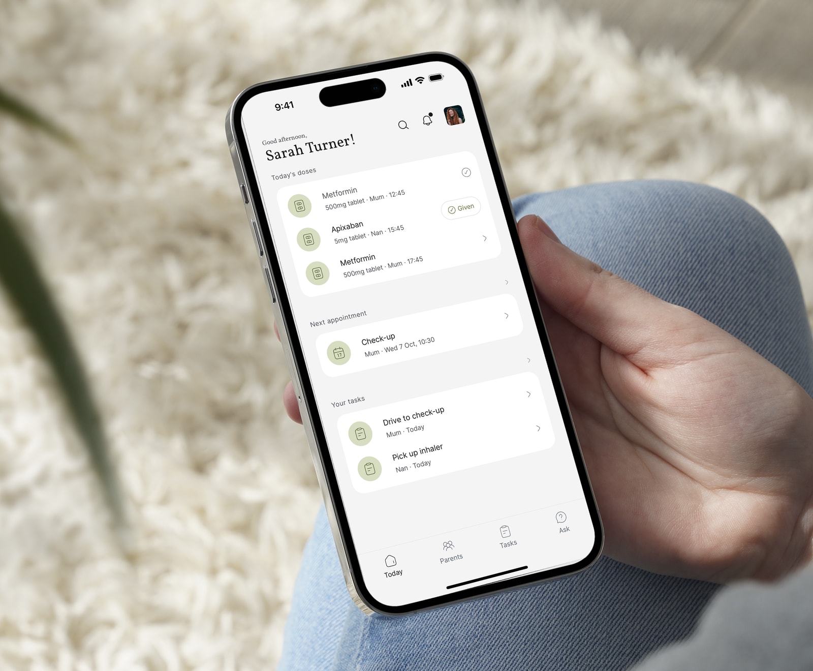 Hand holding an iPhone showing Kinlog's Today screen for Sarah Turner, with Mum's and Nan's doses, Mum's next check-up and the family's tasks