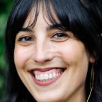 Writer Merav Yami smiles while standing in front of trees and flowers..