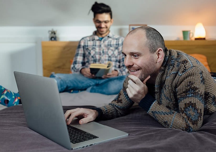 observa-se dois homens na imagem, um sentado em um sofá, e o outro em frente ao sofá, sentado, mexendo no computador