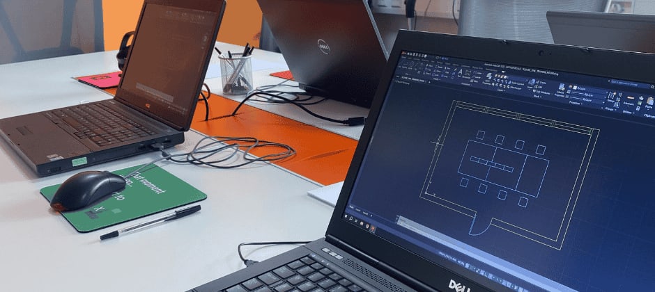 A laptop displaying AutoCAD software on the screen, set up for an AutoCAD training course. The screen shows a technical drawing, with various AutoCAD tools visible in the interface. The laptop is placed on a desk with a notepad and pen nearby.