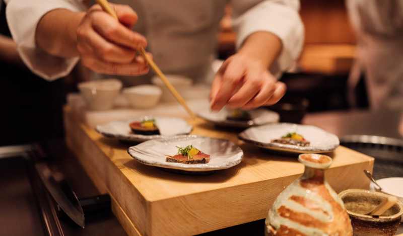 A chef plating delicate courses with chopsticks at an intimate counter, a sake flask in the foreground.