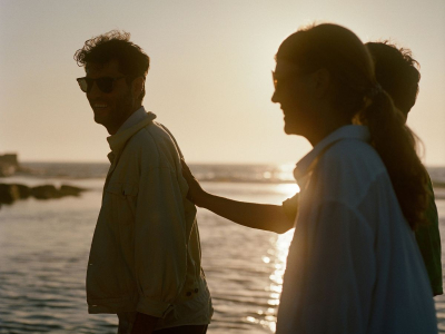 Couple near the ocean at sunset; woman places hand on smiling man's back, both wearing sunglasses, warm golden light.