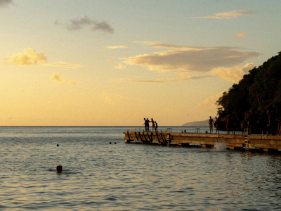 Silhouetted figures on a wooden pier at golden sunset, with calm ocean waters, a lush hillside, and swimmers below.
