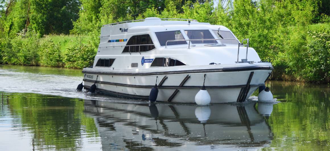 The Clipper cruising slowly along a peaceful tree-lined canal, with bright reflections on the still water ahead.