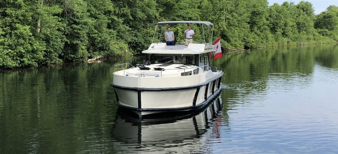 A Horizon boat cruises through a narrow canal surrounded by forest, with a Canadian flag flying from the stern.