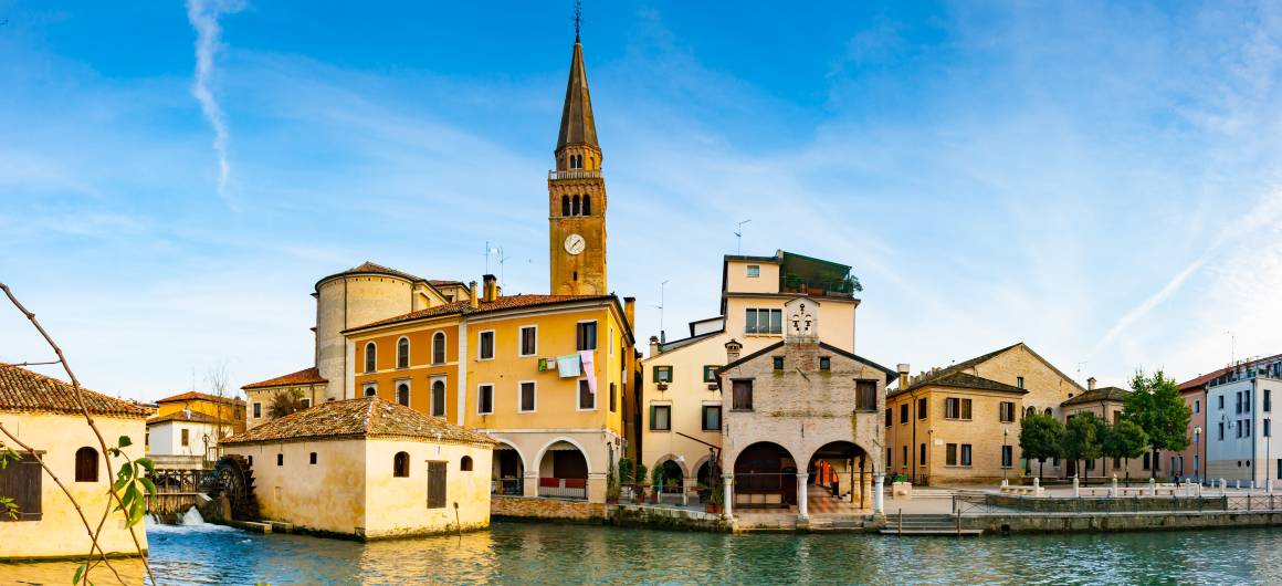 Historic bell tower and yellow‑painted buildings along the riverside in Portogruaro, Veneto.