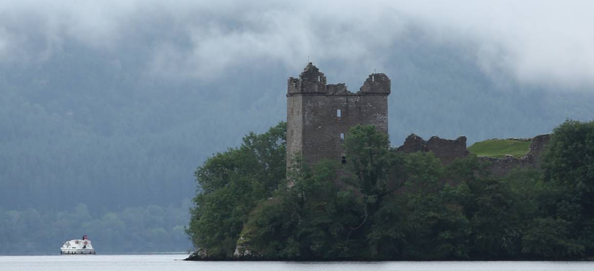 Une vue brumeuse d'une ruine de château sur la rive d'un loch, avec des nuages bas suspendus au-dessus des collines boisées en arrière-plan.