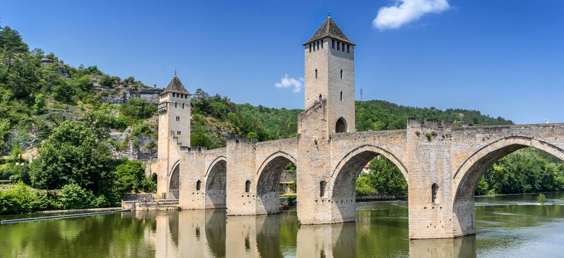 Historic Pont Valentré in Cahors spanning the Lot River, with towers reflected in the calm water.