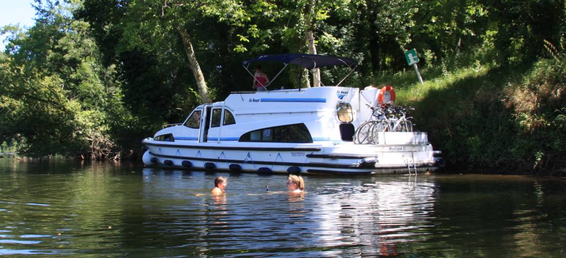 Two people swimming near a moored Le Boat boat on the shaded banks of the Lot River.