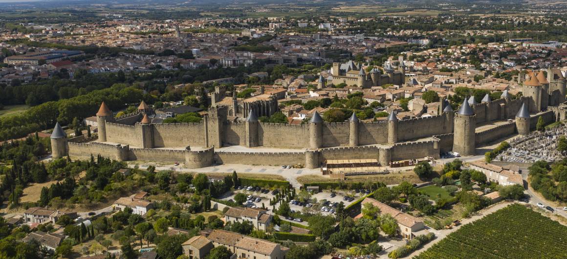 A sweeping aerial view of the medieval citadel of Carcassonne in southern France, with its double-walled fortifications and turrets standing out against the backdrop of the modern city, a must-see landmark for travellers cruising the nearby Canal du Midi.