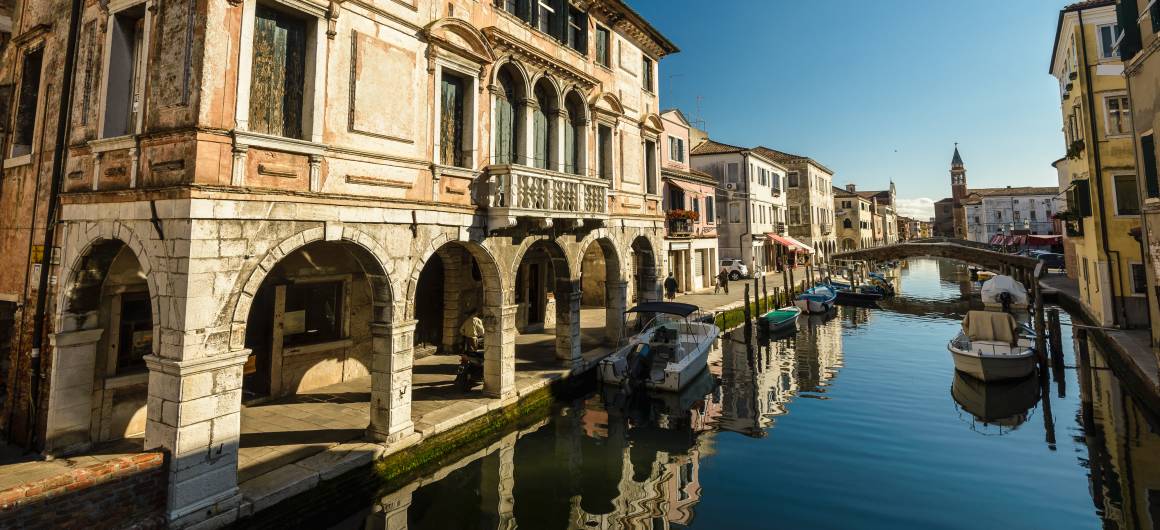 Arched palazzo and moored boats reflected in the canal of Chioggia’s historic centre.