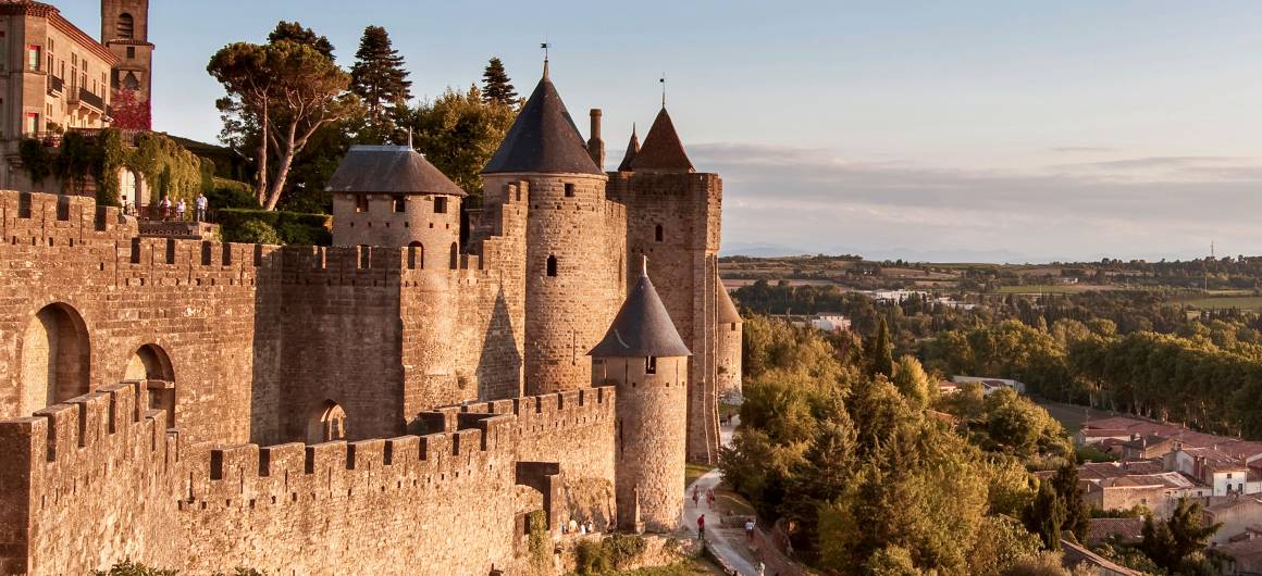 A golden hour view of Carcassonne’s fortified medieval ramparts, with its stone towers and turrets overlooking the peaceful Aude Valley, a highlight of any Canal du Midi cruise in southern France.