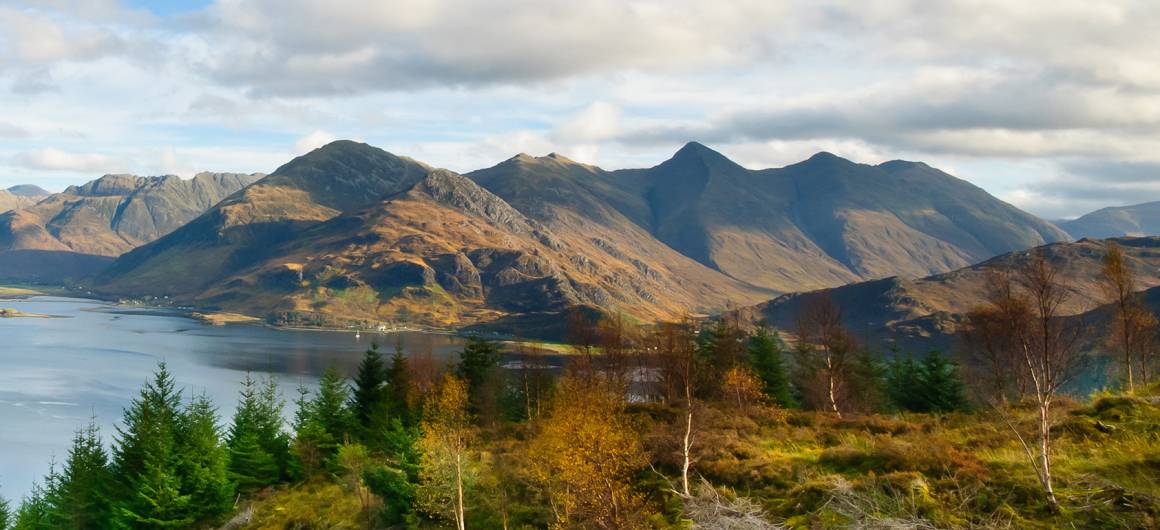 A scenic view of Glen Coe with highland mountains surrounding a tranquil loch and valley, dotted with pine trees and heather.