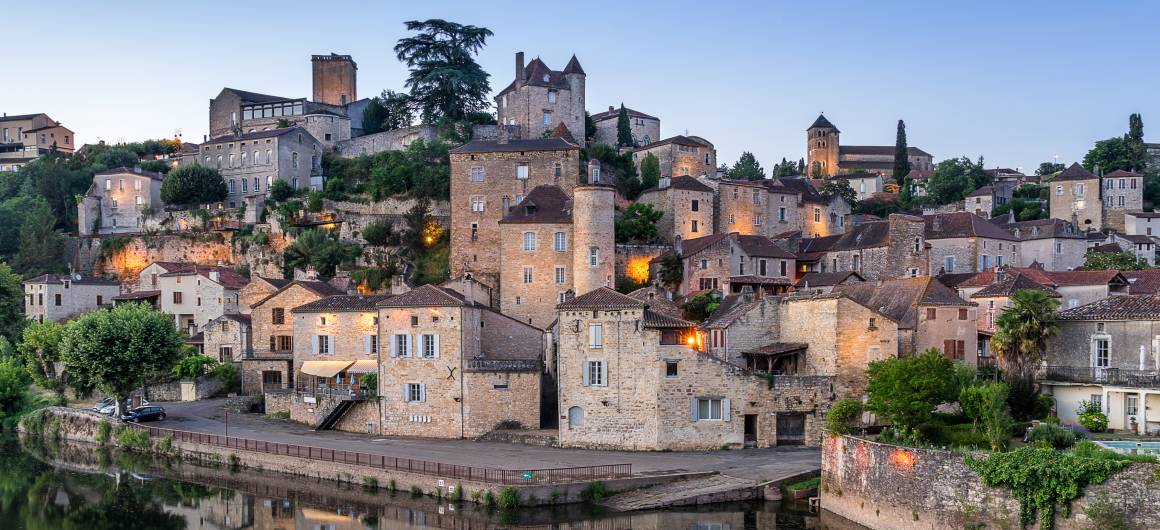 Twilight view of Puy-l’Évêque with stone houses stacked along the riverbank, warmly lit and reflected in the Lot River.