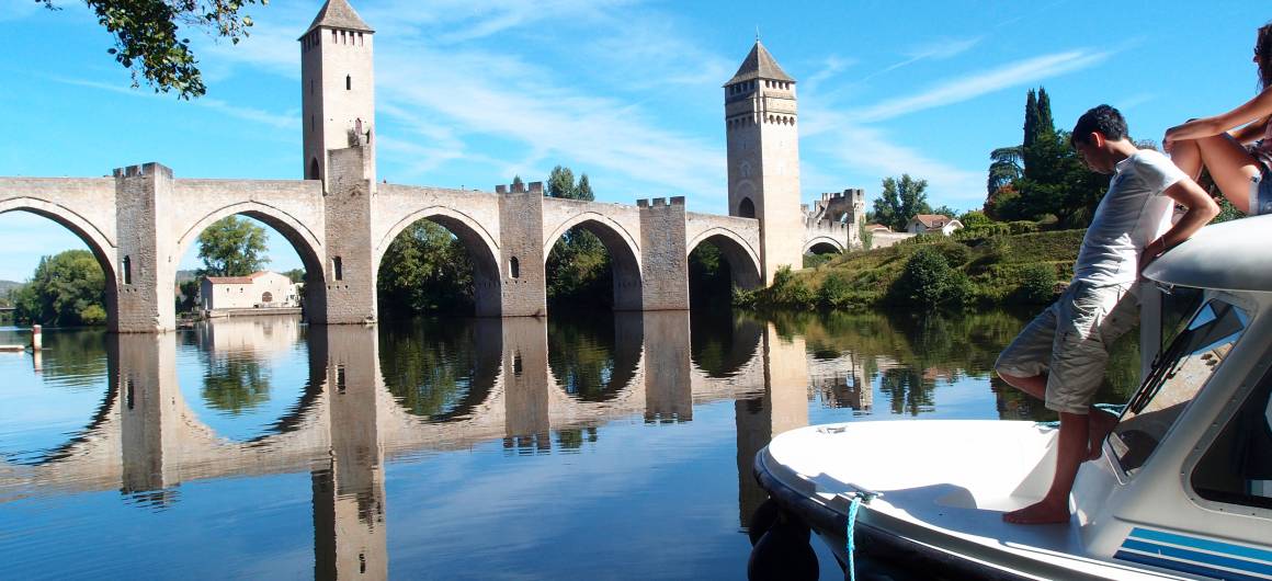 A family aboard a boat admiring the Pont Valentré, perfectly reflected in the mirror-like surface of the Lot River.