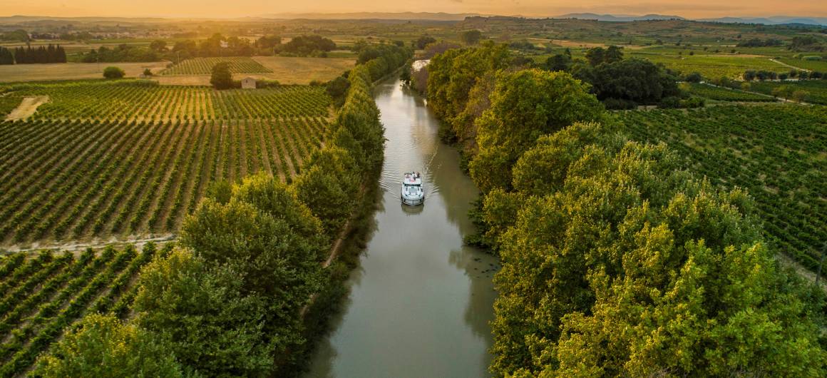 A lone canal boat sails into the golden sunset on the Canal du Midi, cutting through vineyards and rural countryside for a picture-perfect moment on a French boating holiday.