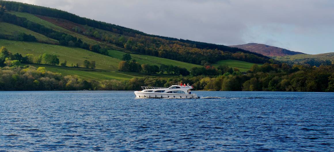 A modern white cruiser sailing across a loch, backed by patchworked green and forested hills under a sunny sky.