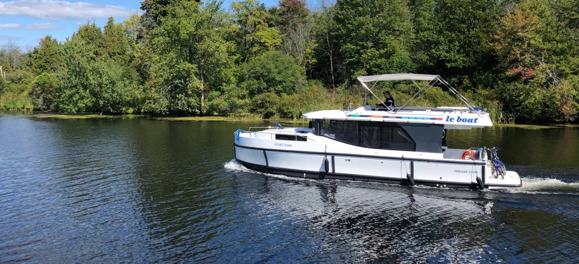 A Horizon boat glides through a quiet stretch of the Rideau Canal, framed by lush green trees under a bright blue sky.