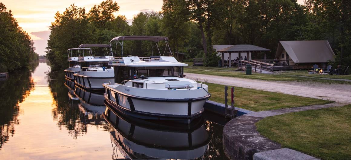 Several Horizon boats are moored along the Rideau Canal beside a small campground under a pink sunset sky.