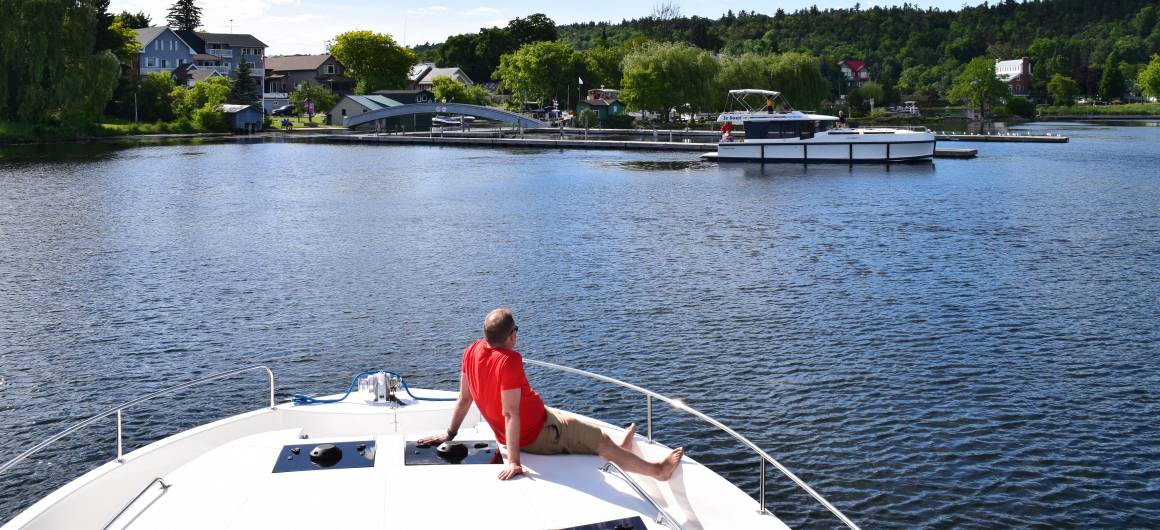 A man relaxes on the bow of a Le Boat boat approaching a riverside town along the Rideau Canal on a sunny day.