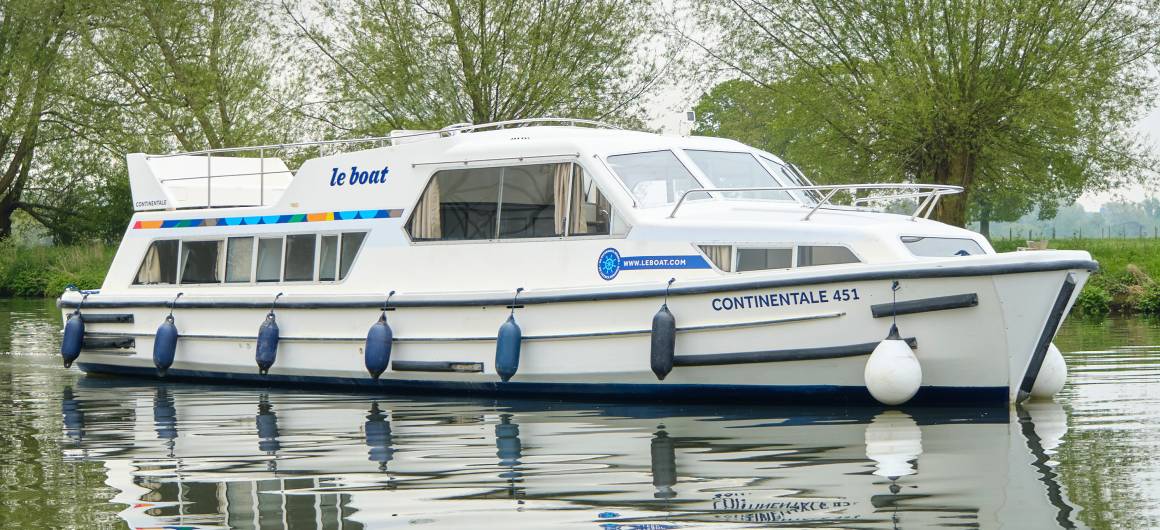 Side view of the Continentale gliding across a peaceful canal, reflected clearly in the still water and set against a backdrop of spring greenery.