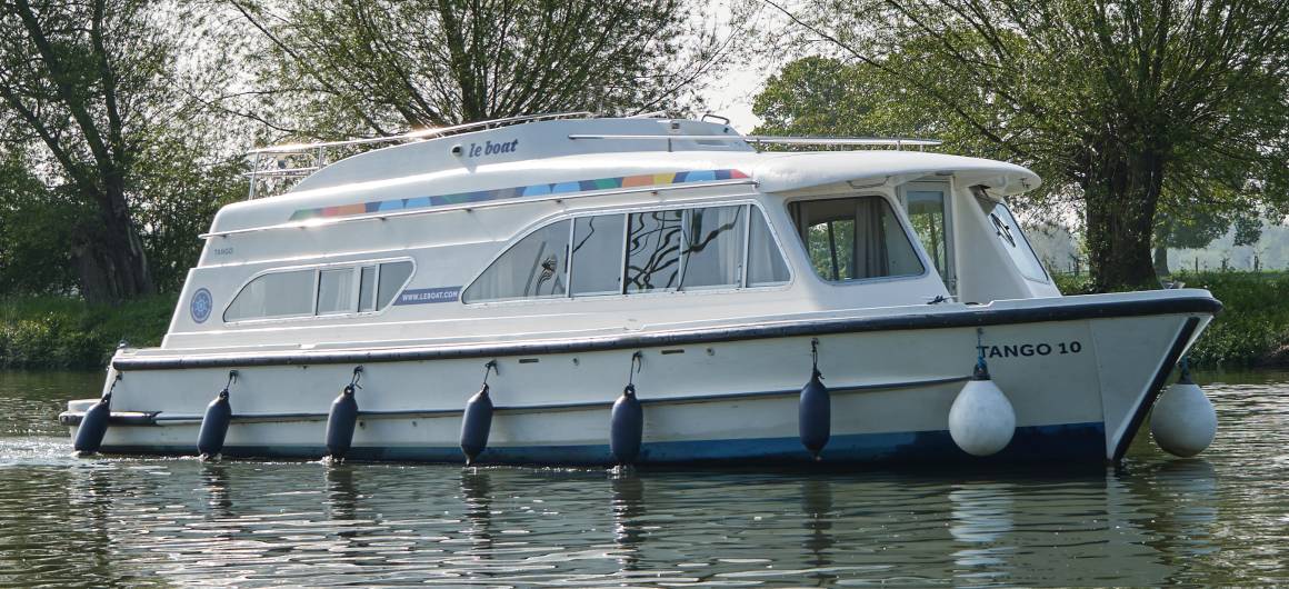 Dancer 2 cruising gently along a calm river, framed by green trees and reflected in the glassy surface of the water under a clear blue sky.