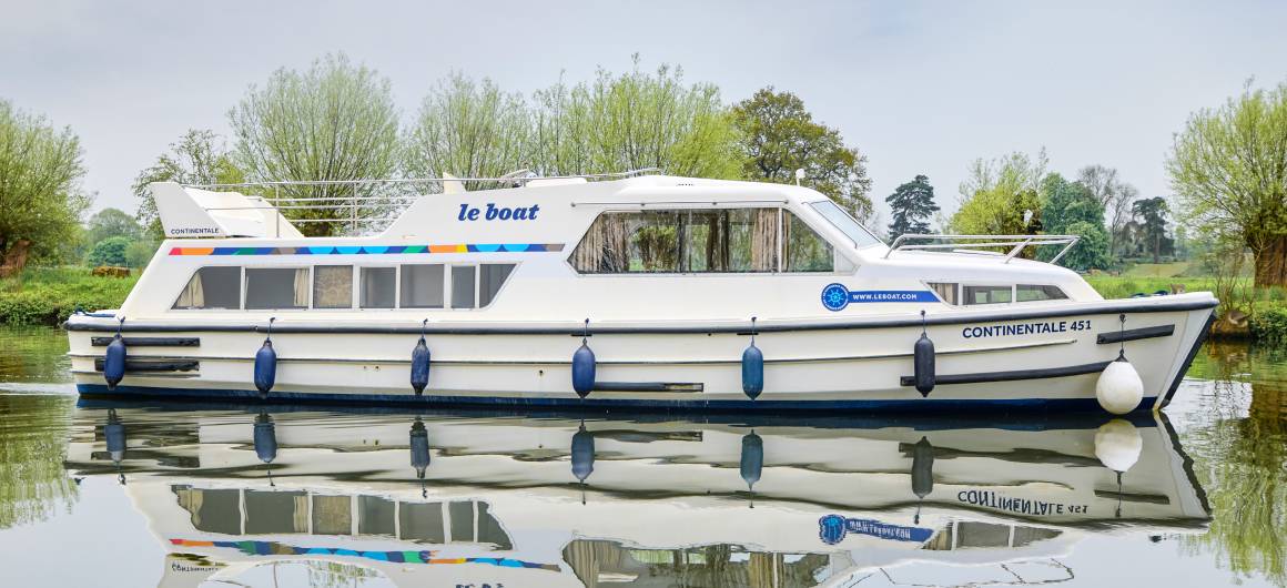 Side view of the Continentale cruising gently along a quiet river, framed by trees and mirrored beautifully in the calm, glassy water.
