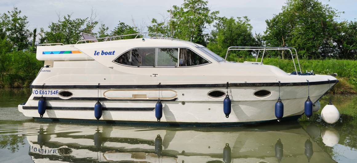 Side view of the Countess on a quiet canal, its sleek profile reflected in the still water beneath a bright blue sky.
