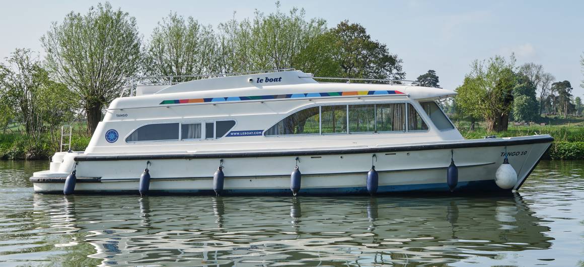 The Dancer 2 glides smoothly past overhanging trees on a tranquil stretch of river, its streamlined design and shaded windows reflected on the water.