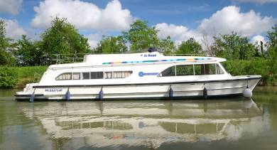 Side view of the Dancer 4 cruising along a peaceful canal, reflected clearly in the calm green water.