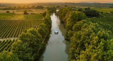 A lone canal boat sails into the golden sunset on the Canal du Midi, cutting through vineyards and rural countryside for a picture-perfect moment on a French boating holiday.