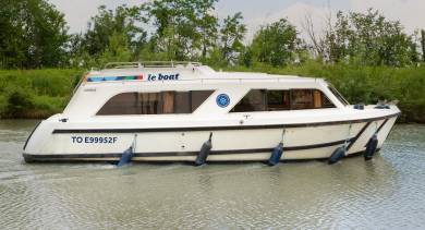 Side view of the Cirrus B gently moving along the canal, with still water and a hedge-lined towpath under a blue sky.
