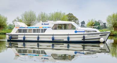 Side view of the Continentale cruising gently along a quiet river, framed by trees and mirrored beautifully in the calm, glassy water.