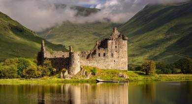 Die Ruinen von Kilchurn Castle spiegeln sich im ruhigen Wasser des Loch Awe, umgeben von nebligen grünen Hügeln in Argyll, Schottland.
