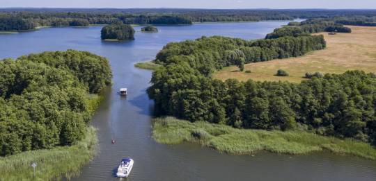 Two boats cruise through a lush, tree-lined channel linking Germany’s interconnected lakes under a clear summer sky.
