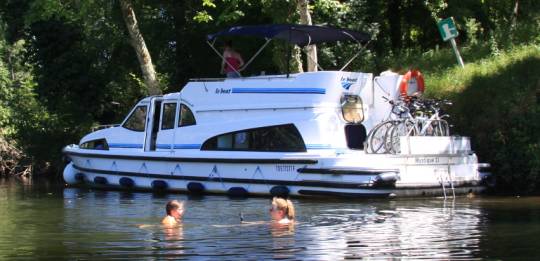 Two people swimming near a moored Le Boat boat on the shaded banks of the Lot River.
