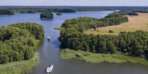 Zwei Boote fahren durch einen kanalartigen Wasserweg, gesäumt von Bäumen, der Deutschlands verbundene Seen unter einem klaren Sommerhimmel verbindet.
