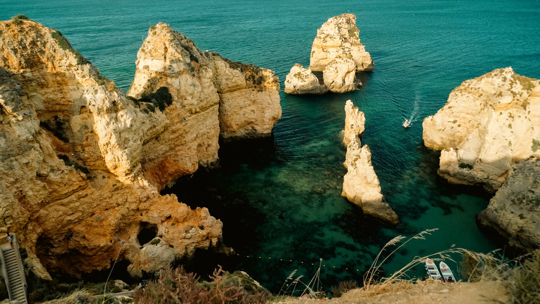 Aerial view of the dramatic Algarve coastline in Portugal, featuring golden limestone sea stacks and rock formations rising from clear turquoise-green water. A natural rock arch is visible on the left, with a sheltered cove below. Small boats are moored near the base of the cliffs, and another boat leaves a wake in the open water in the background.