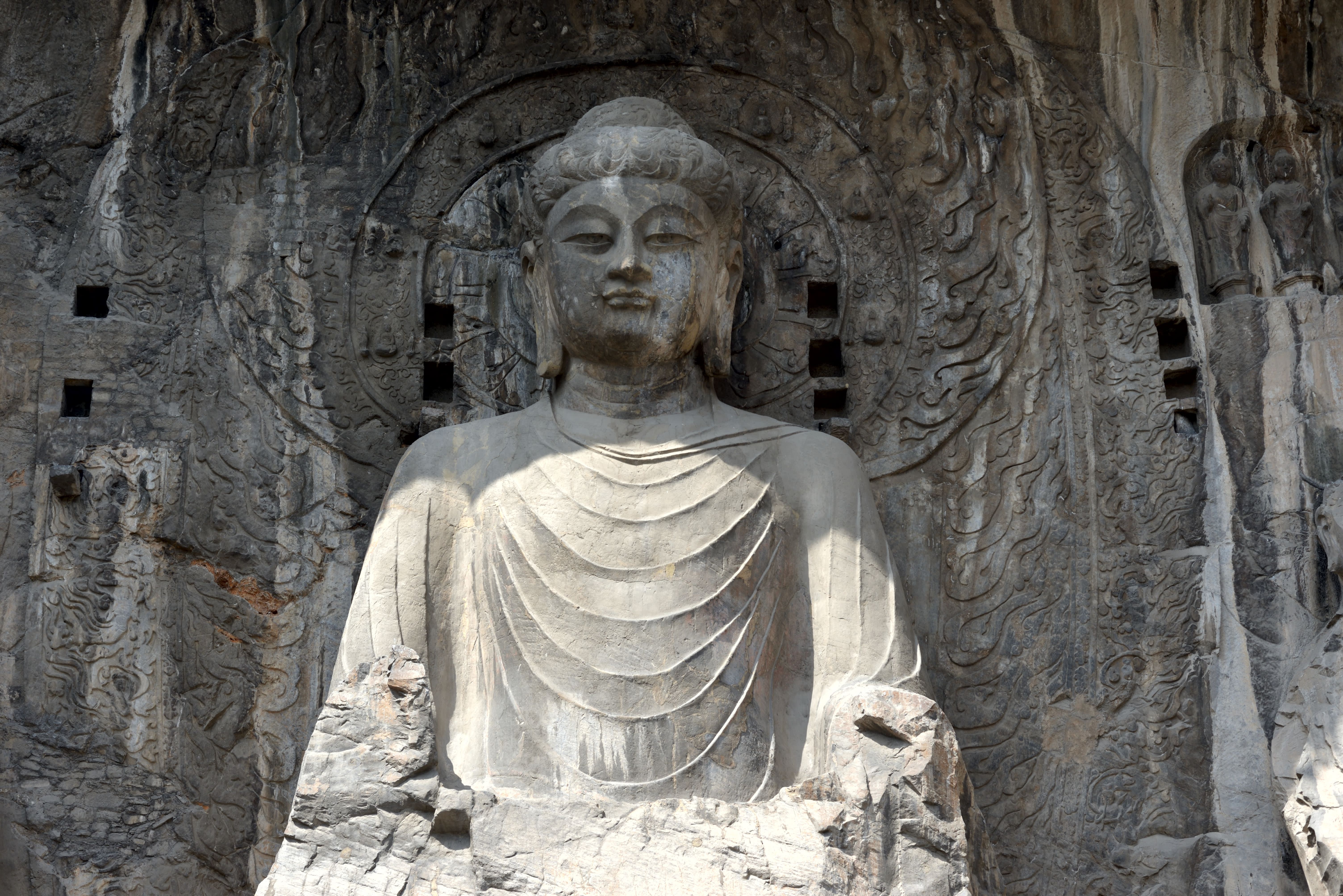 Large ancient Buddha statue carved into a rock cliff, with a circular halo and flowing robes, surrounded by intricate stone reliefs.