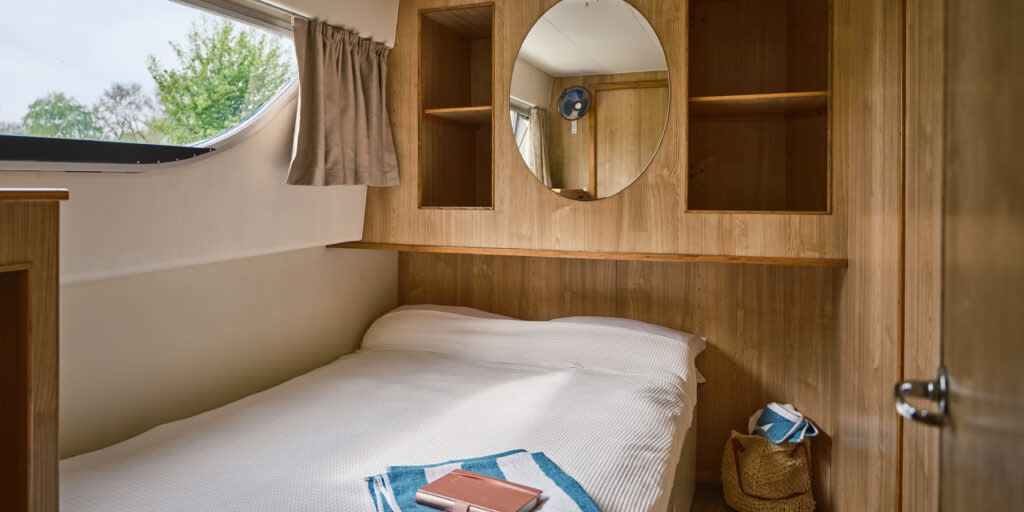 Comfortable double cabin on the Dancer 3 featuring a round wall mirror, built-in shelving, and large window offering views of the riverside greenery.