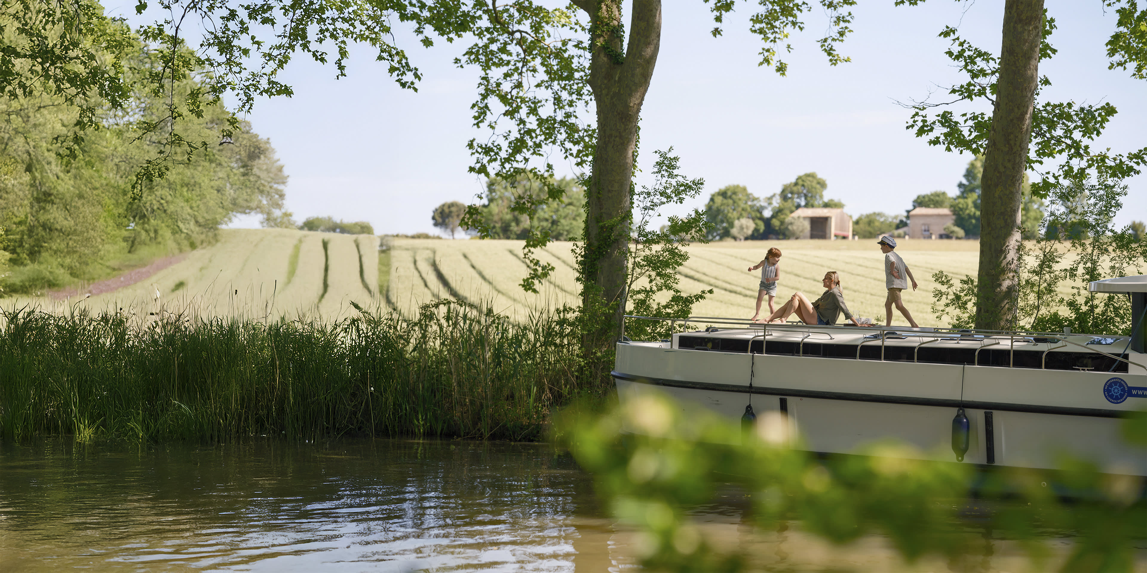 Zwei Kinder spielen, während ihre Mutter sich auf dem Boot entspannt und einen sonnigen Tag am Flussufer genießt.