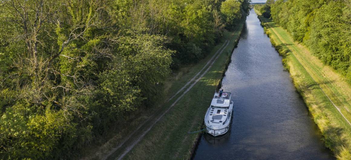 Vue par drone d’un bateau de plaisance naviguant entre forêt et lac sur le passage étroit du canal de Réchicourt.