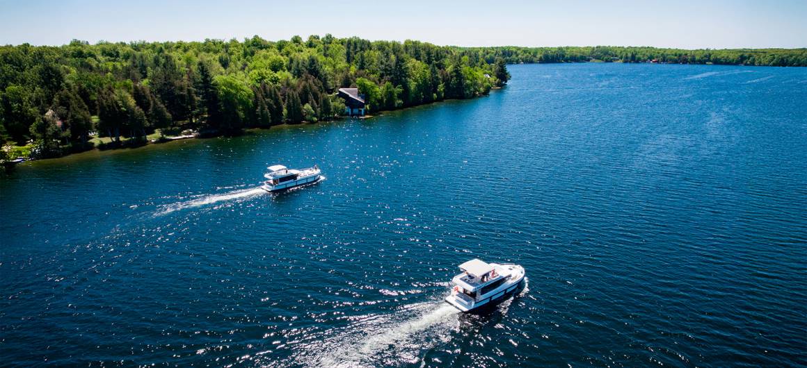 Two Horizon boats cruise across sparkling blue waters along a tree-lined stretch of the Rideau Canal in Ontario, Canada.