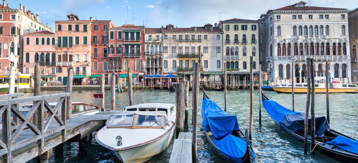 Moored boats and gondolas in front of elegant historic buildings along the Grand Canal in Venice.