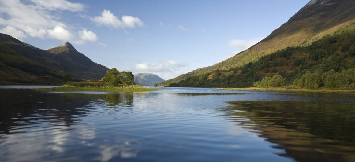 Un large loch bordé de montagnes des Highlands et de pentes boisées, avec des nuages reflétés doucement dans l’eau calme.