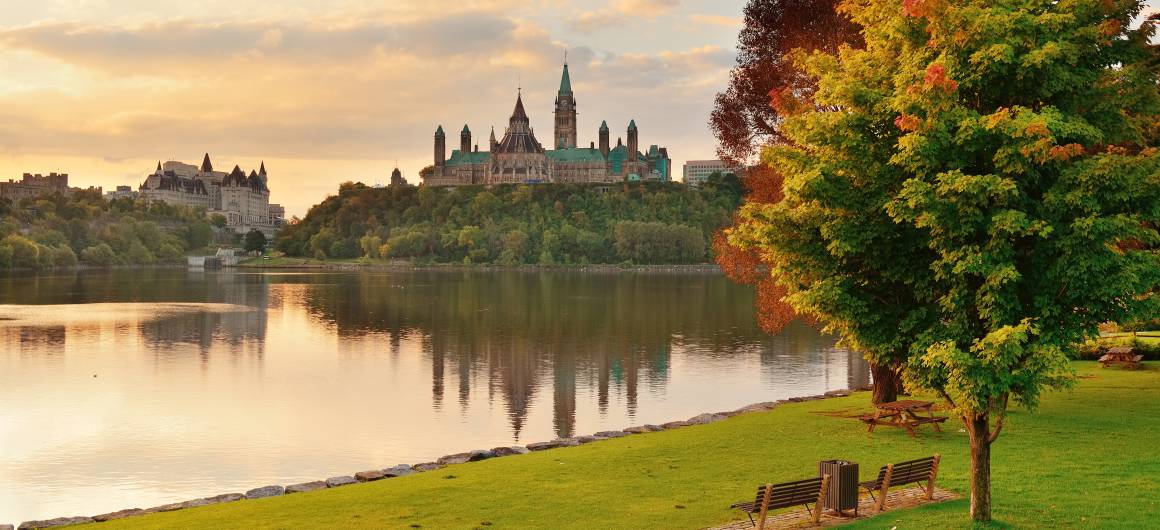 View across the Ottawa River with Parliament Hill and Château Laurier lit by morning light, seen from a park across the water.
