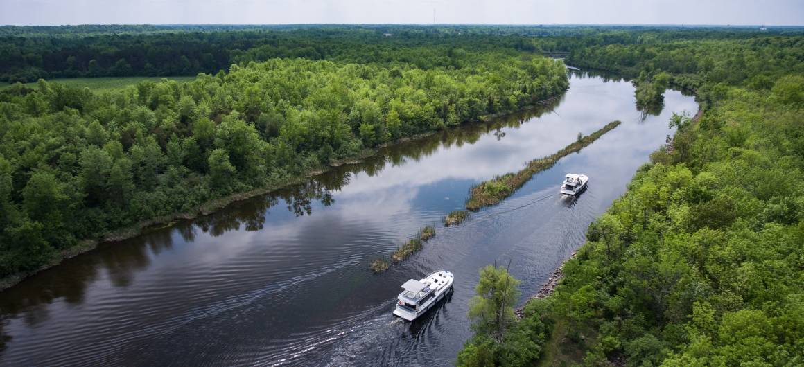 Two Le Boat boats navigate a calm, tree-lined channel of the Rideau Canal, their wake trailing behind in the reflective water.