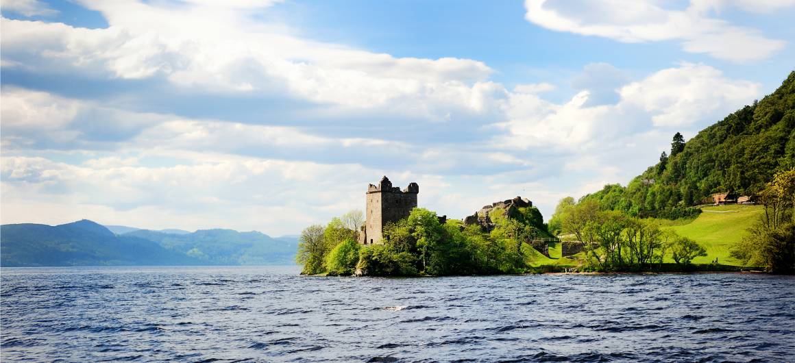 Un château en pierre partiellement intact se dresse au bord du Loch Ness, entouré d'arbres verts luxuriants et de collines herbeuses.