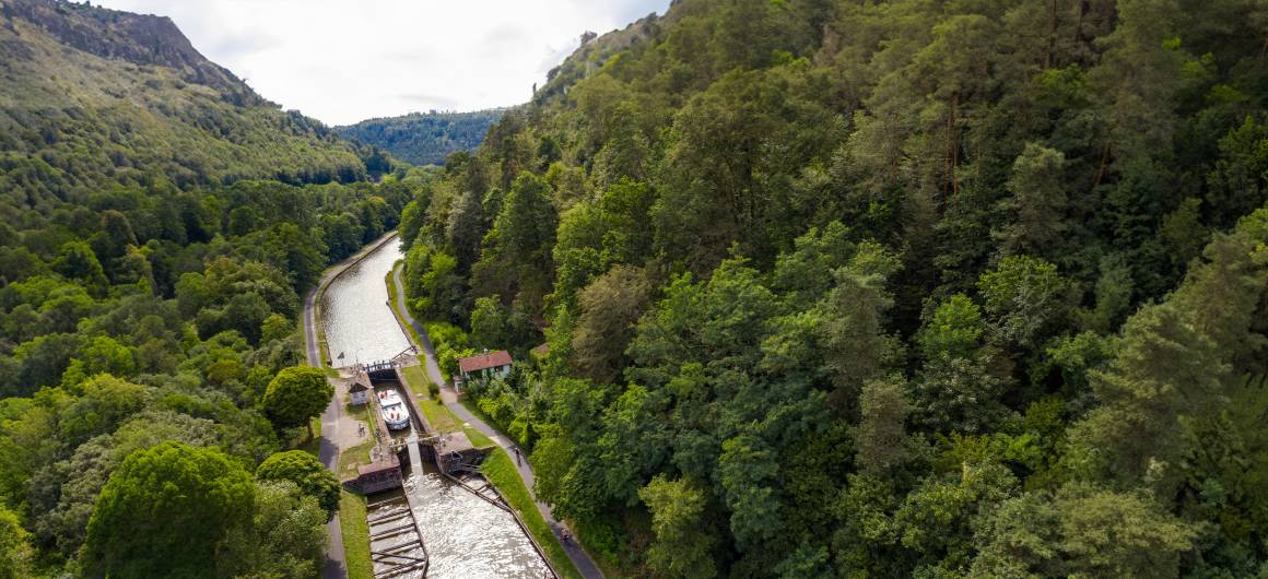 Bateau entrant dans une écluse nichée dans une vallée forestière luxuriante au pied des Vosges.