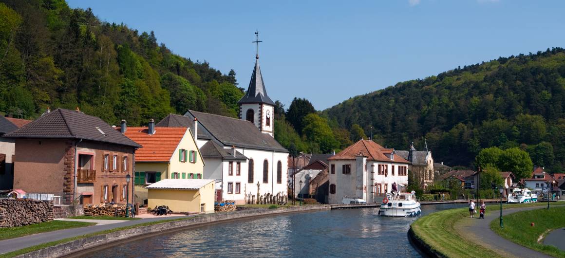 Bateaux circulant sur le canal dans un village de la Hesse, avec des maisons colorées, une église et des collines verdoyantes sous un ciel lumineux.